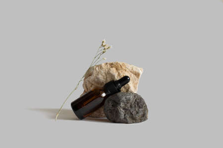 Brown dropper glass pipette bottle laying on rocks with flower branch, gray background, copy space. Serum, acid on podium. Zero waste glass. Organic, natural cosmetic. Spa, beauty, skincare productの写真素材