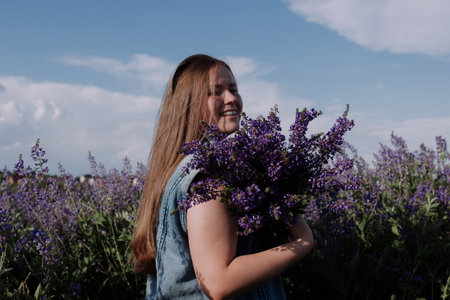 Smiling young woman in denim jacket holding bouquet of purple flowers standing alone among field in front of blue sky enjoying of sunlight at golden hour. Summer outfit. Girl with flowersの写真素材