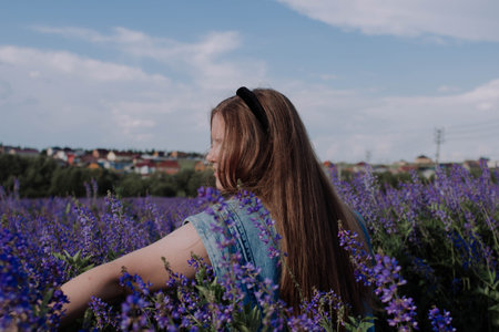 Smiling young woman in denim jacket touching with hand purple flowers standing alone among field...