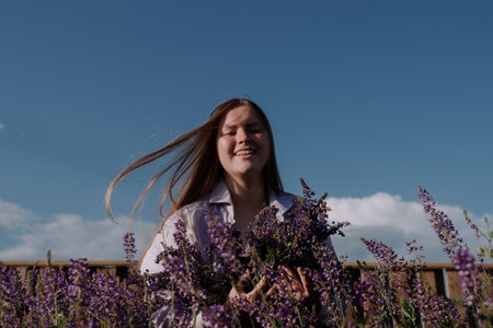 Smiling young woman in shirt holding bouquet of purple flowers standing alone among field in front wooden fence and blue sky enjoying of sunlight at golden hour. Summer outfit. Girl with flowersの写真素材