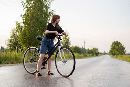 Young woman standing alone on road with bicycle at countryside. Summer activity, healthy lifestyle,...