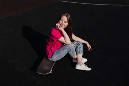 Young woman in pink t-shirt with closed eyes sitting on skateboard outside. Summer activity, healthy...
