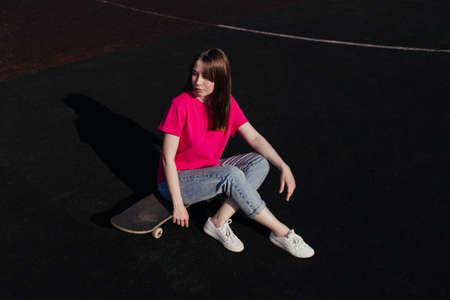 Young woman in pink t-shirt sitting on skateboard outside looking to the right. Summer activity, healthy lifestyle, workout, sport, fitness, riding. Female skater. Having fun outdoors.の写真素材