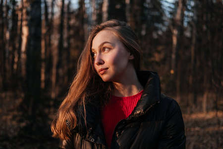 Smiling young woman in coat and scarf standing in autumn forest. Woman looking at the sunset on...