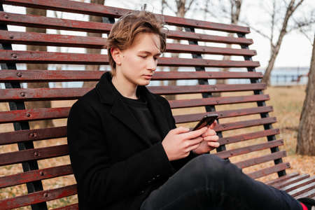 Young man with birthmarks, moles, wart on face in black coat with smartphone in hands sitting alone on wooden bench on a street. Working on mobile device out of home outdoors. Freelancer, bloggerの写真素材