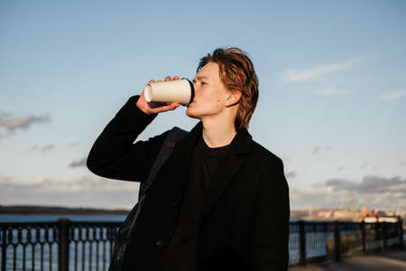Handsome young man in black coat drinking coffee to go and walking on a city street in the morning. Blonde man having a break with hot beverage, relaxing in autumn park. Fatigue after work.の写真素材