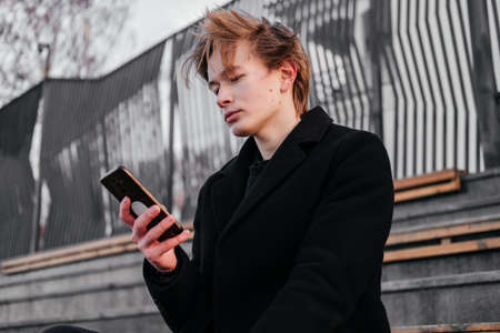 Young man with birthmarks, moles, wart on face in black coat with smartphone in hands sitting alone...