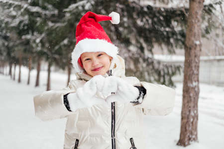 Laughing little girl in red christmas hat standing under snowfall and doing hand heart in mittens....