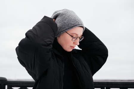 Pensive young woman with short hair in hat, eyeglasses, black coat with hands up touching head....
