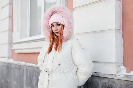 Ginger woman in pink coat hood standing on a street in front of building wall looking at camera. Redhead girl 20 years old outdoorsの写真素材