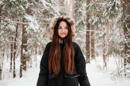 Young woman in black coat walking in winter forest and smiling. Girl with long hair in park in front of trees outdoorsの写真素材