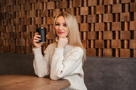 Smiling mid age woman holding cup of coffee sitting at cafe indoors. Caucasian girl in white sweater.の写真素材