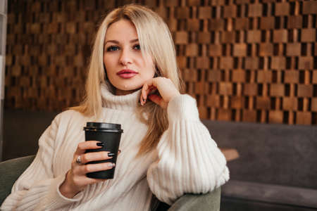 Mid age woman drinking coffee at cafe looking at camera. Caucasian blonde girl in white sweater indoors.の写真素材