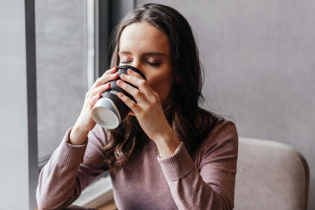 Caucasian young woman drinking coffee sitting at cafe indoors. Woman enjoying coffee beverageの写真素材