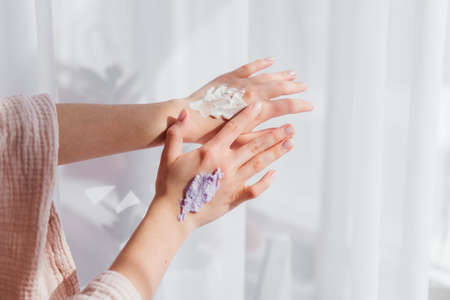 Female hands applying purple cream scrub on skin at home side view white background. Skincare routine conceptの写真素材