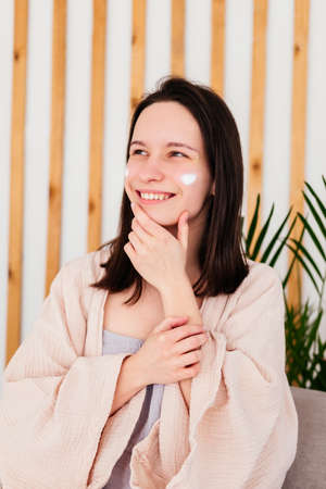 Caucasian young woman touching face and applying cream indoors. Morning beauty routine. Skincare...