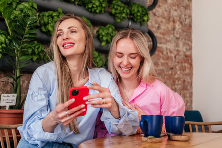 Two women friends sitting in cafe drinking coffee and laughing. Smiling caucasian girls chatting indoorsの写真素材