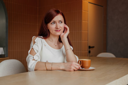 Smiling mid age woman with red hair hair sitting in cafe and drinking coffee looking away indoors.の写真素材