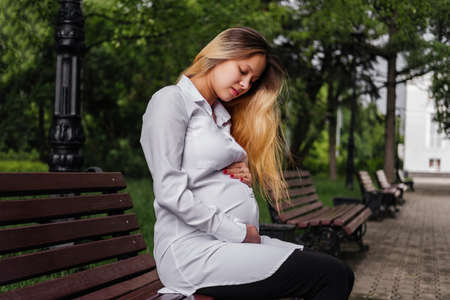 Pregnant young woman sitting on bench and holding hand on belly outdoorsの写真素材