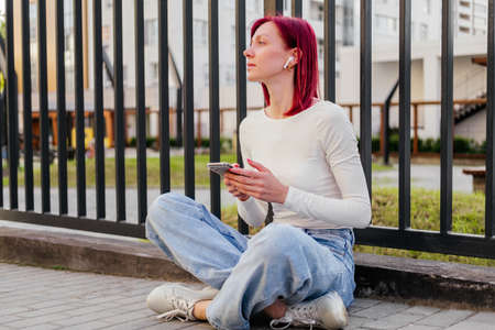 Young woman in earphones sitting on the ground with smartphone outdoor. Girl listening music in the cityの写真素材