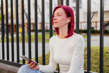 Woman using mobile phone listening music in earphones sitting on ground outdoor. Girl with closed eyes relaxing in cityの写真素材