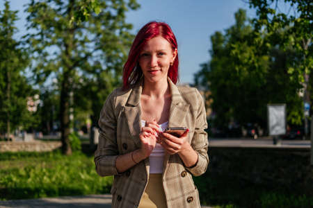 Smiling woman with red hair standing with smartphone in hands outdoor. Girl using smartphone looking at cameraの写真素材