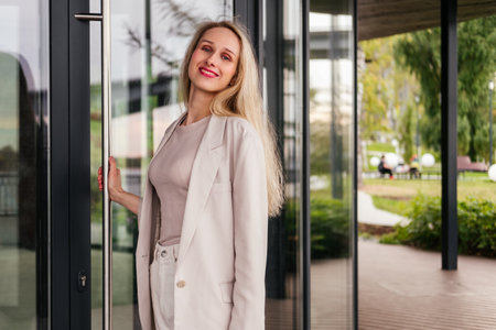 Smiling woman standing near office building and opening door. Middle aged businesswoman in white clothes outdoorの写真素材