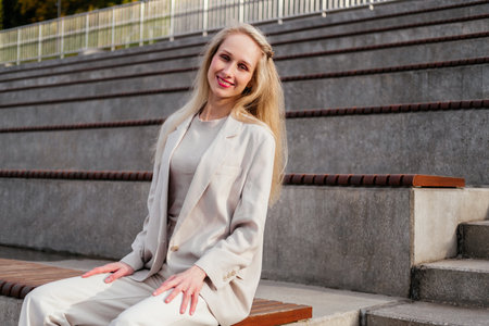 Smiling middle aged businesswoman in white clothes sitting on bench in park looking at camera outdoorの写真素材