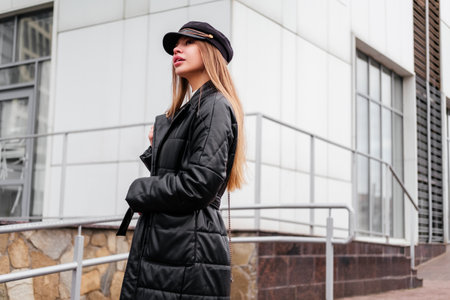 Elegant young woman in black coat and beret standing near office building looking away outdoorの写真素材