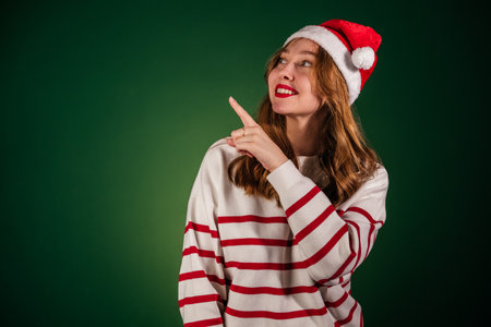 Woman in santa hat holding new year gift isolated on green background. Excited girl in sweater pointing index finger aside. New year and christmas celebration conceptの写真素材
