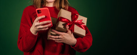 Female hands using smartphone and holding new year gift isolated on green background copy space. Unrecognizable woman in sweater with boxes. online shopping conceptの写真素材