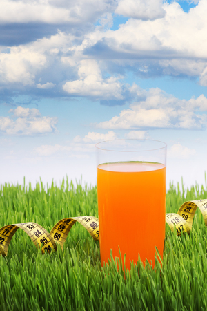 A glass of orange juice on the green grass, the meter coiled, glass of juice on a background of the cloudy sky, a healthy diet, the young wheat, diet and sport lifestyle.の写真素材