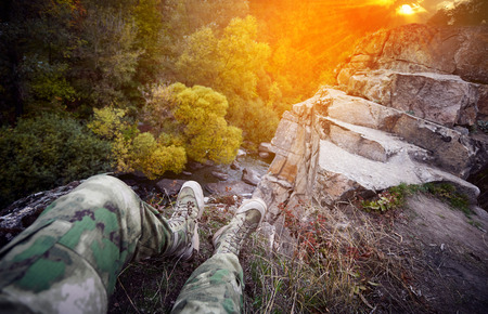 Traveler at a mountain river, legs over a cliff, outdoor clothing, deep stone canyon, ancient granite rocks, beautiful sunset, rocky river bank, cloudy sky, mountain landscape, forest on the riverbank, sun rays.の写真素材