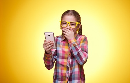 Emotional teenager, the little girl plays with the phone, children's emotions, girl with glasses, bright checkered shirt, beautiful young lady, portrait of a young girlの写真素材