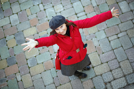 Photo of a smiling woman in a red jacket and a gray beret on a background of old paving slabs. Model posing arms to the side and looking up at the camera. Shallow depth of field. Focus on the modelの写真素材