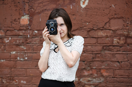Girl with an old vintage camera on a brick wall background. Young woman photographer taking photo outdoor. Selective focus on model. Retro style photo.の写真素材