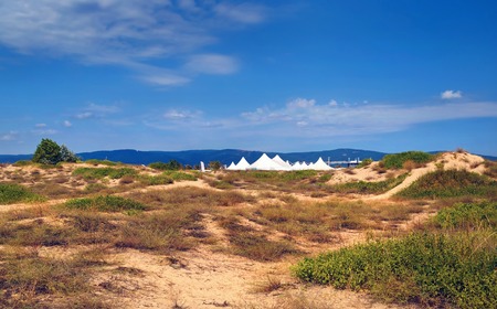 Summer sunny day. Blue sky and dry plants on the sand. White tents and mountains in the background. Black Sea coast.の写真素材