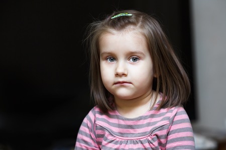Close-up portrait of a serious little girl against dark background. Child looks into the camera.の写真素材
