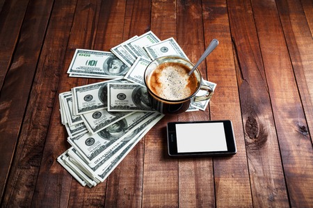 Coffee cup, money, dollars and phone on vintage wooden table background. Business concept.の写真素材