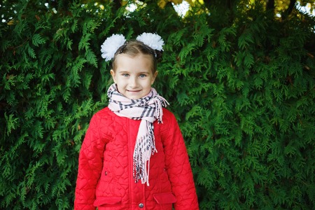 Smiling baby girl in a red jacket with white bows on her head. Child posing on a background of green...