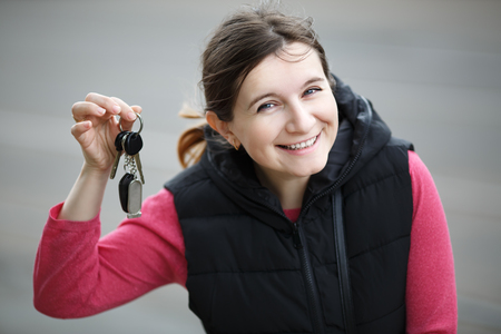 Smiling young woman holding car key on blurred gray asphalt background. Selective focus.の写真素材
