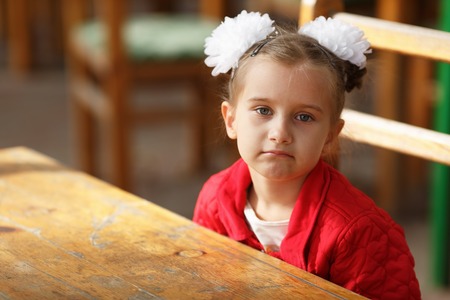 Portrait of a little girl in a red jacket with white bows on her head. Child sitting at the table. Shallow depth of field. Selective focus.の写真素材