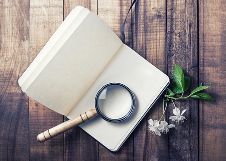 Still life with a notepad. Blank opened notebook, magnifier, cherry flowers and green leaves on vintage wood background. Stationery elements. Responsive design mockup.の写真素材