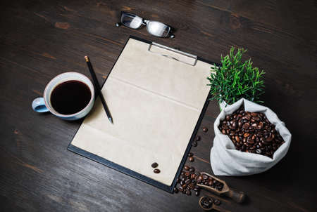 Coffee and blank menu. Clipboard with blank kraft letterhead, coffee cup, coffee beans, plant, glasses and pencil on wood table background.の写真素材