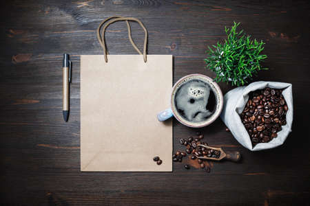 Blank kraft paper bag, coffee cup, plant, coffee beans in canvas bag and pen on wooden background....