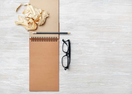 Opened notebook with blank craft paper pages, glasses, pencil and crumpled paper on light wood table...
