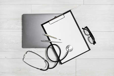 Various medical supplies. Blank clipboard, stethoscope, glasses, thermometer, pen and laptop on wooden background. Flat lay.の写真素材