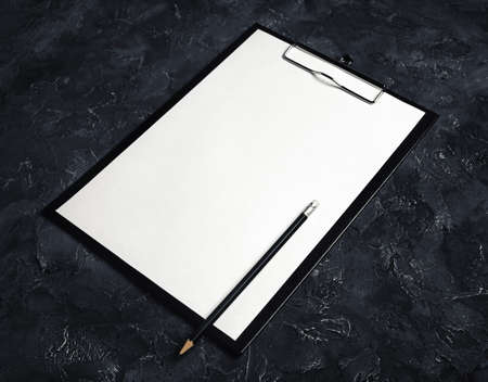 Photo of clipboard with a blank sheet of paper and pencil on black plaster background. Mockup for branding identity.の写真素材