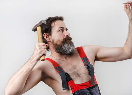 Bearded builder in overalls hammers a nail with a hammer. Man with hammer.の写真素材