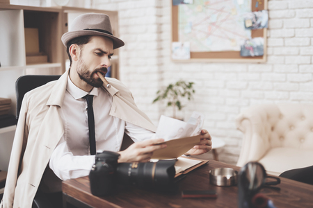 Private detective agency. Man in hat and cloak is sitting at desk smoking sigar and looking at photos.の写真素材
